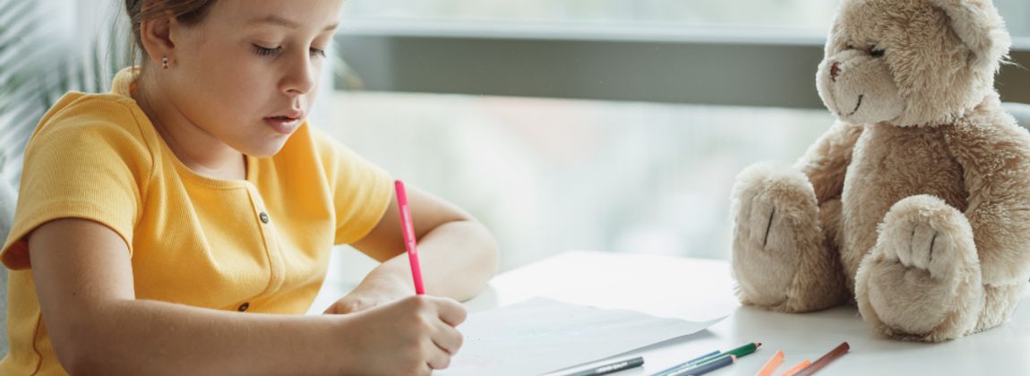 A child writing at a desk beside a soft toy