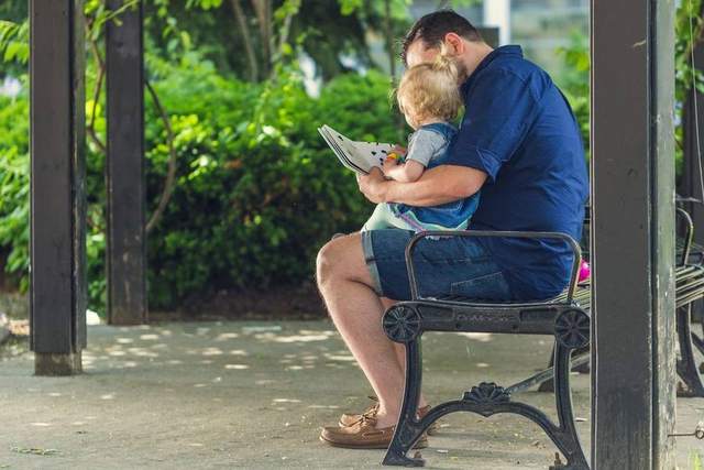 An adult and a small child sitting together on a park bench, sharing a picture book, seen from behind
