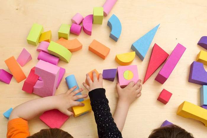 Brightly coloured foam blocks in geometric shapes spread across a wooden table, with two children reaching in to build with them