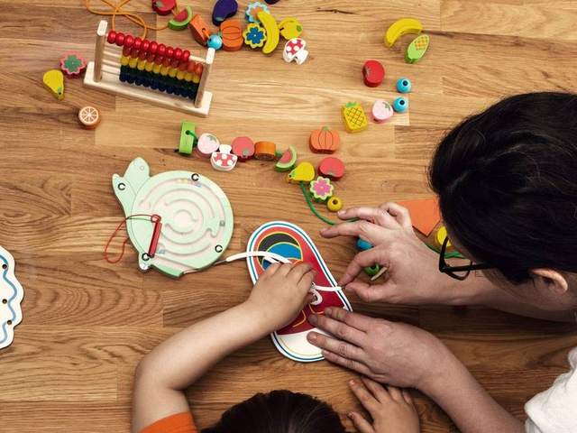 An adult sitting with a small child on the floor, helping them thread a lace through a wooden activity card