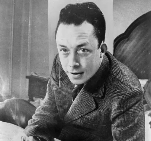 Albert Camus with dark swept-back hair in a checked tweed jacket and tie, leaning forward on a desk and looking at the camera in a 1957 black-and-white press photograph