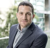 Andrew Huberman, a neuroscientist, in a gray blazer over a black-and-white checked shirt, smiling slightly against a blurred green and glass background