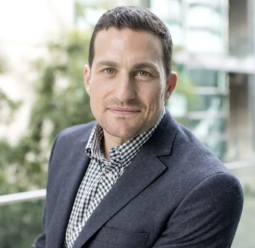 Andrew Huberman, a neuroscientist, in a gray blazer over a black-and-white checked shirt, smiling slightly against a blurred green and glass background