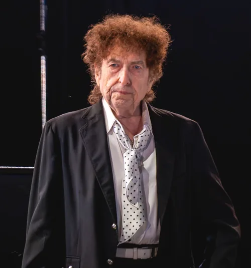 Bob Dylan with curly auburn hair, in a dark jacket over a white shirt and a polka-dot neckerchief, standing on stage during a concert in Kilkenny, Ireland, in July 2019