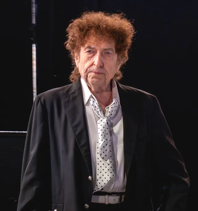 Bob Dylan with curly auburn hair, in a dark jacket over a white shirt and a polka-dot neckerchief, standing on stage during a concert in Kilkenny, Ireland, in July 2019