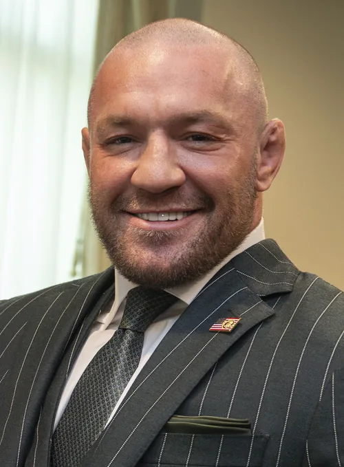 Conor McGregor with a shaved head and a broad smile in a grey pinstripe suit in March 2025