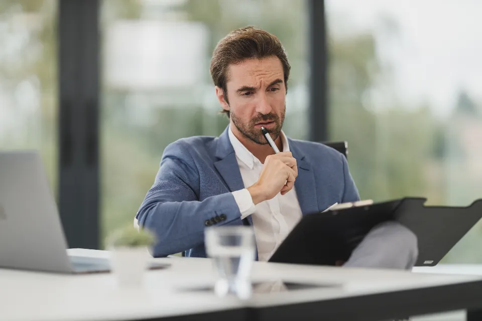 A man in an office thinking something over, pen held to his lips