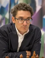 Fabiano Caruana, a chess grandmaster with curly brown hair and dark-framed glasses, in a dark blazer over a white shirt, leaning over the board with chess pieces in the foreground, in January 2025