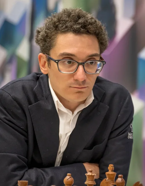 Fabiano Caruana, a chess grandmaster with curly brown hair and dark-framed glasses, in a dark blazer over a white shirt, leaning over the board with chess pieces in the foreground, in January 2025