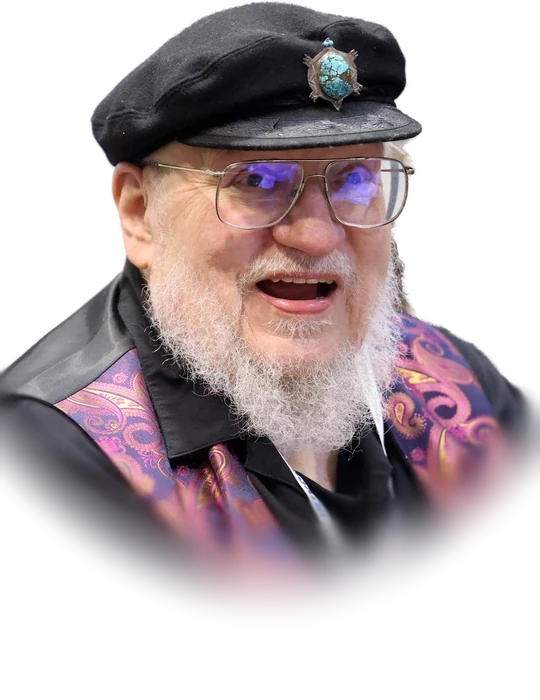 George R. R. Martin in a black captain’s cap and glasses, with a white beard and a paisley-patterned vest, smiling mid-sentence in August 2025