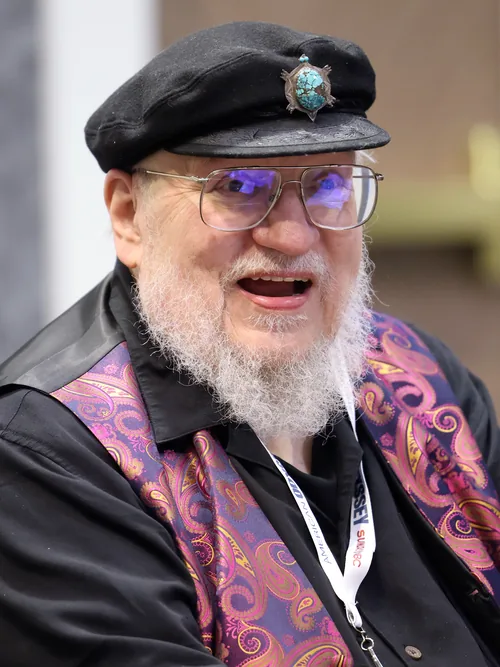George R. R. Martin in a black captain’s cap and glasses, with a white beard and a paisley-patterned vest, smiling mid-sentence in August 2025