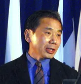 Haruki Murakami in a dark suit, blue shirt and striped tie, speaking at a podium in 2009