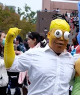 A cosplayer in yellow face paint with large googly eyes and a white dress shirt dressed as Homer Simpson, with a raised yellow-gloved fist, at Comic World Taiwan in December 2015