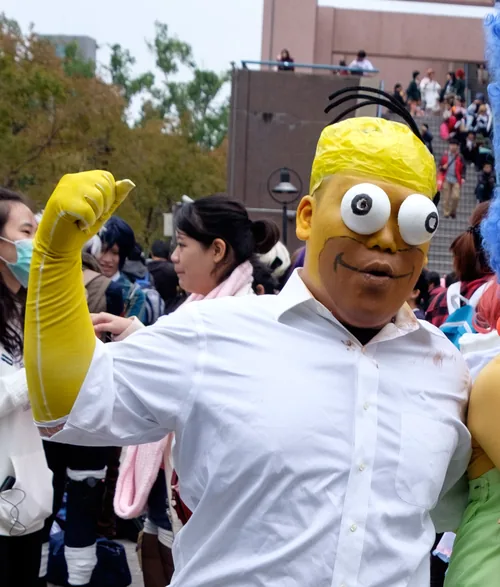 A cosplayer in yellow face paint with large googly eyes and a white dress shirt dressed as Homer Simpson, with a raised yellow-gloved fist, at Comic World Taiwan in December 2015