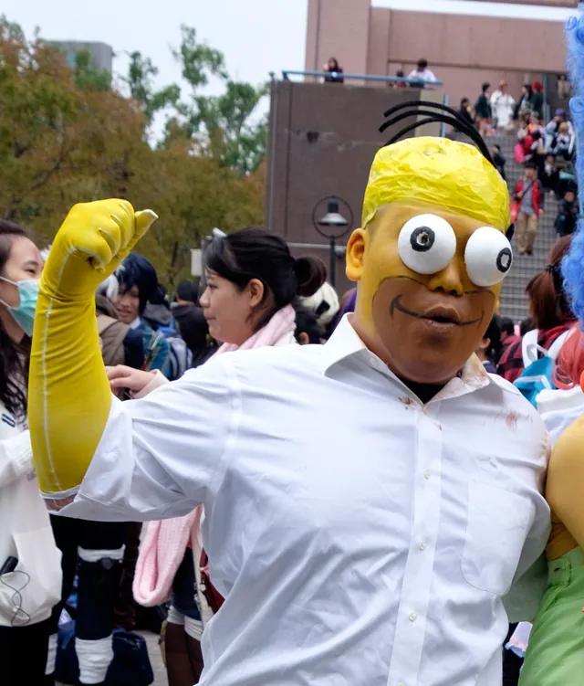 A cosplayer in yellow face paint with large googly eyes and a white dress shirt dressed as Homer Simpson, with a raised yellow-gloved fist, at Comic World Taiwan in December 2015