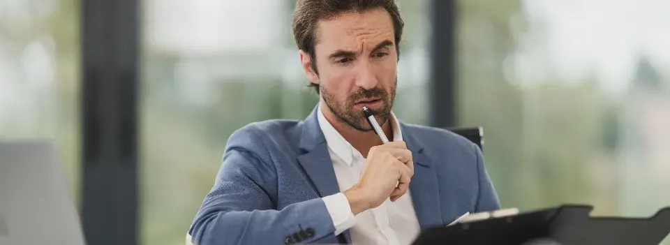 A man in an office thinking something over, pen held to his lips