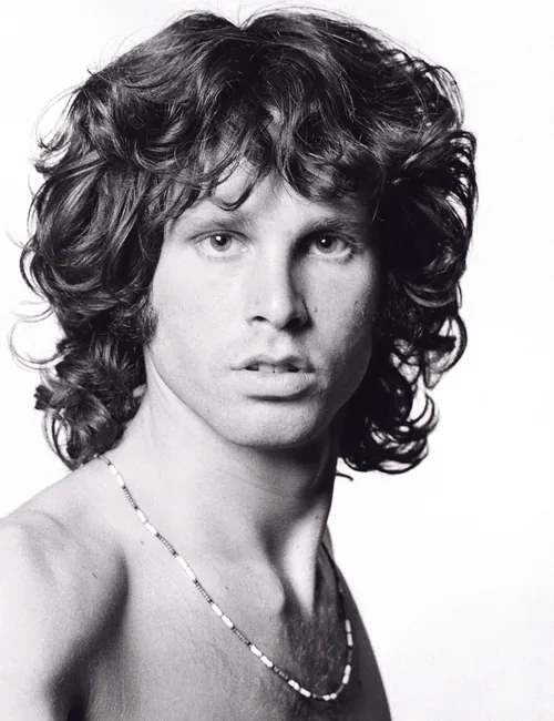 Jim Morrison with tousled dark curly hair looking at the camera in a black-and-white 1967 studio portrait