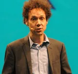 Malcolm Gladwell with curly reddish-brown hair in a brown jacket and a light-blue open-collared shirt, speaking on stage in November 2011