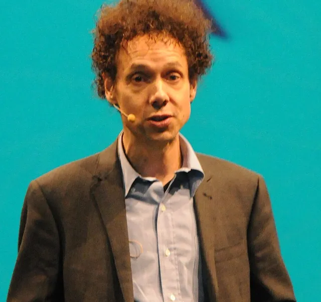 Malcolm Gladwell with curly reddish-brown hair in a brown jacket and a light-blue open-collared shirt, speaking on stage in November 2011