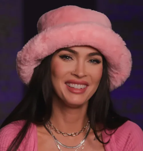Megan Fox smiling in a pink fur bucket hat and a pink cardigan in 2023