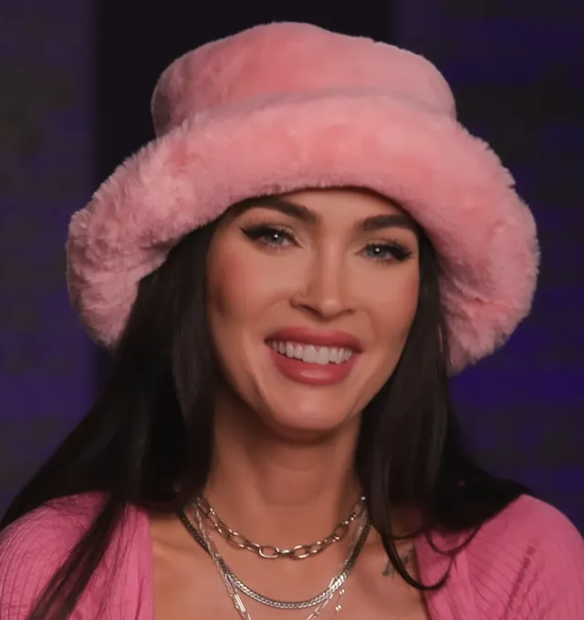 Megan Fox smiling in a pink fur bucket hat and a pink cardigan in 2023