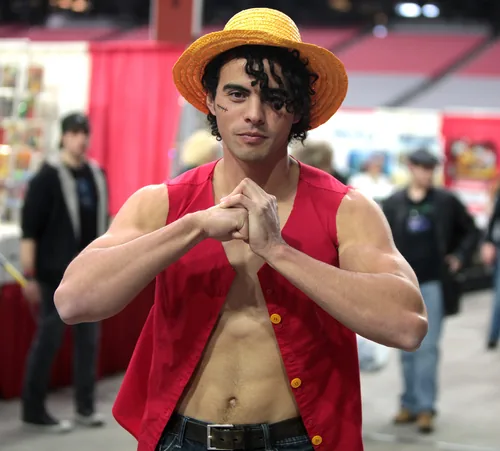 A cosplayer dressed as Monkey D. Luffy from One Piece, in a straw hat and an open red vest, with both hands pressed together in front of the chest, photographed at a convention in December 2015