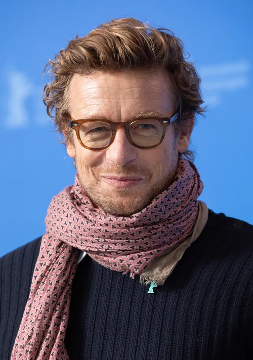 Simon Baker, the actor who played Patrick Jane in The Mentalist, smiling in tortoiseshell glasses and a pink patterned scarf at the Berlin International Film Festival in February 2020