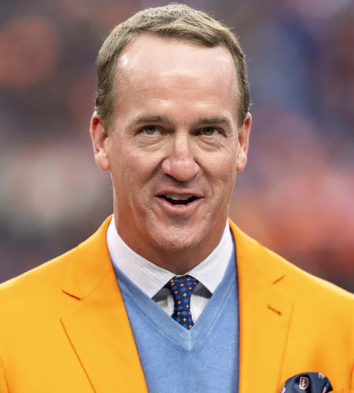 Peyton Manning in an orange blazer and a blue sweater speaking at a Denver Broncos game in October 2021