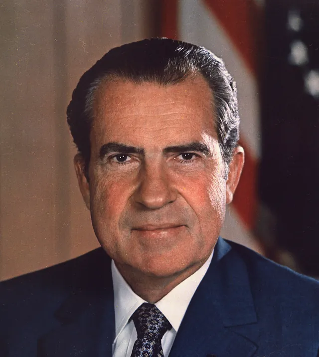 Richard Nixon in a dark blue suit and patterned tie with a small American flag pin on the lapel, in his official presidential portrait from about 1972