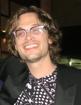 Matthew Gray Gubler, the actor who played Spencer Reid in Criminal Minds, smiling in clear-framed glasses with wavy light-brown hair, a black jacket and a black-and-white patterned shirt, in New York City in October 2017