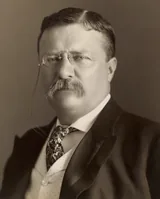 Theodore Roosevelt in pince-nez glasses with a mustache, a dark jacket, waistcoat and patterned tie, in a sepia studio photograph from 1904
